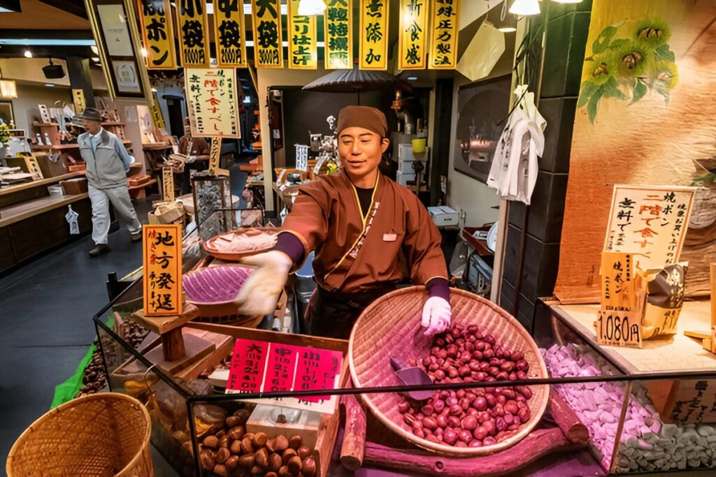 Navigationpaths.com | Kyoto Travel Secrets: Hidden Temples, Sacred Rituals & Undying Charm A vendor in traditional clothing sells chestnuts at a colorful Japanese market stall, surrounded by baskets of produce and signs with Japanese writing. Another person is visible in the background.