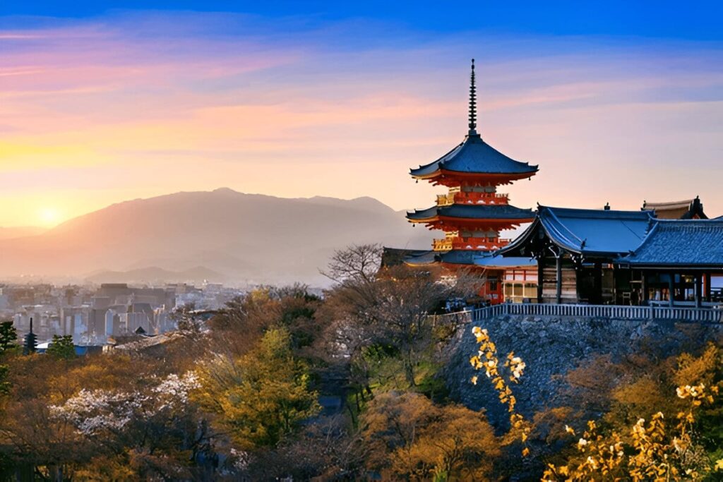 Navigationpaths.com | Kyoto Travel Secrets: Hidden Temples, Sacred Rituals & Undying Charm A traditional Japanese pagoda and temple sit atop a hill surrounded by trees with autumn foliage, overlooking a cityscape at sunset with mountains in the background.